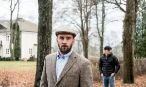 Man med skägg och tweedkavaj bär en klassisk gubbkeps engelska. I bakgrunden syns ett hus och en annan man.
