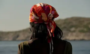 En kvinna med färgglad bandana i håret, en stil som visar hur knyter man en bandana. Bakgrunden är en lugn sjö och berg.