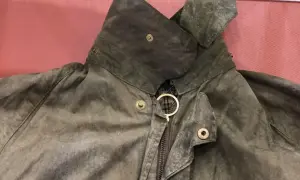 Detaljerad bild av en sliten Barbour-jacka, redo att laga. Kragen är uppfälld och visar dragkedjan med en gyllene ring.