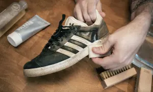 Händer rengör en smutsig Adidas Gazelle-sneaker med en trasa. Bredvid ligger rengöringspasta och borstar, redo att impregnera sneakers.