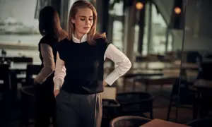 En kvinna med blont hår och fräknar bär en vit skjorta och en svart väst. Hon visar hur en frack ska sitta, med en avslappnad men elegant hållning.