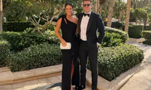 Ett par klädda för black tie-tema, hon i en elegant svart klänning, han i smoking. De poserar glatt utomhus bland grönska.