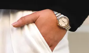 Elegant märkesklocka dam med gulddetaljer på ett silverfärgat armband. Klockan sitter på en handled som sticker ut ur en svart tröja och vita byxor.