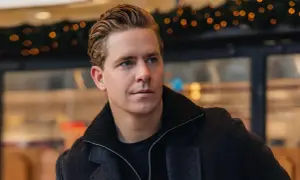 Martin Hansson i stilren höstmode, med en mörk rock och uppsatt hår. Bakgrunden har julbelysning.