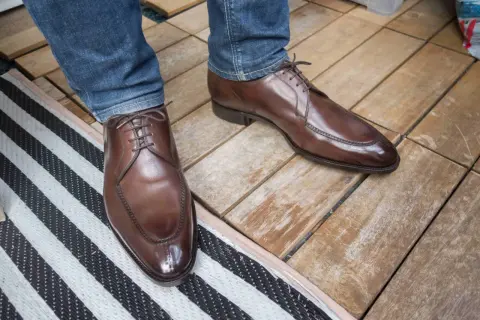 Ett par eleganta bruna läderskor med snörning, som sitter på en person med blå jeans. Skorna har en fin glans och ser ut att vara av hög kvalitet, trots att de har lite glappande skor-känsla.