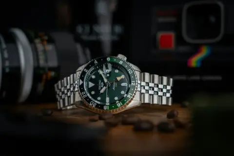 En grön Seiko GMT-klocka med en tvåfärgad bezel, omgiven av kaffebönor. Bakgrunden visar en kamera.
