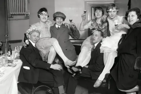 Festligt sällskap i mode 1920-tal, med kvinnor i klänningar och män i kostym, skålar och njuter av festen.