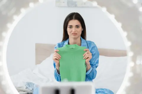 En kvinna i blå pyjamas håller upp en grön topp och ser bekymrad ut, som om hon upptäckt att bomullen krymper.