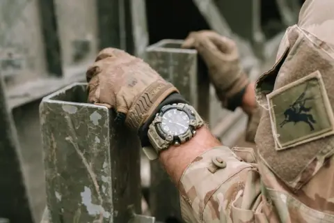 En soldat med moderna klockor på handleden, klädd i kamouflageuniform, håller i en metallkonstruktion.