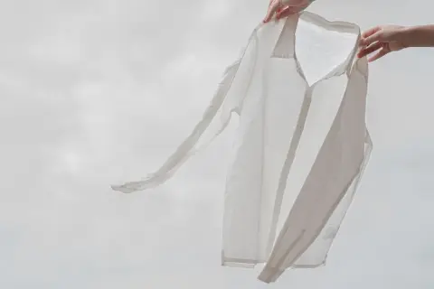 Två händer håller upp en vit, lätt skjorta i viskosmaterial mot en molnig himmel. Skjortan fladdrar i vinden.
