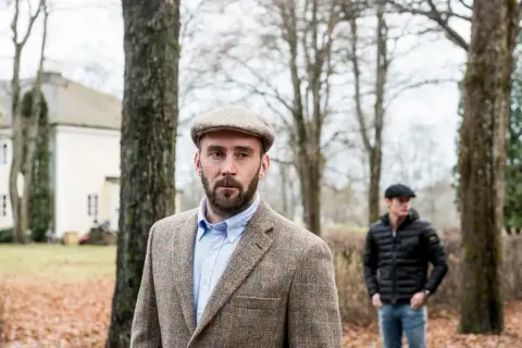 Man med skägg och tweedkavaj bär en klassisk gubbkeps engelska. I bakgrunden syns ett hus och en annan man.