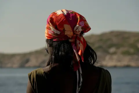 En kvinna med färgglad bandana i håret, en stil som visar hur knyter man en bandana. Bakgrunden är en lugn sjö och berg.