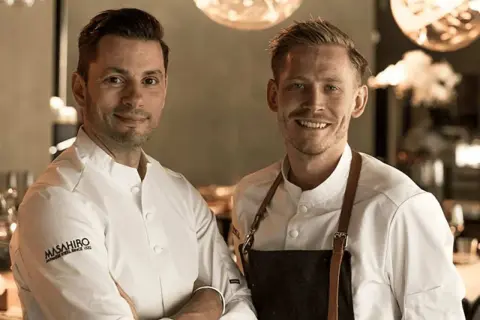 Två kockar ler mot kameran. Deras vita kockrockar och förkläden antyder en professionell miljö, kanske en restaurang som får bra allegrine recensioner.