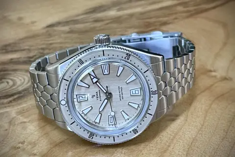 En Yema Superman dykarklocka med silverfärgad urtavla och ett unikt, bikakeformat metallarmband ligger på ett träbord.