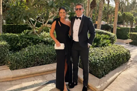 Ett par klädda för black tie-tema, hon i en elegant svart klänning, han i smoking. De poserar glatt utomhus bland grönska.