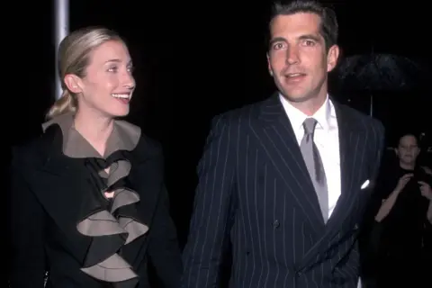 Carolyn Bessette Kennedy och John F. Kennedy Jr. på en tillställning. Hon bär en svart kavaj med volanger, han en randig kostym.