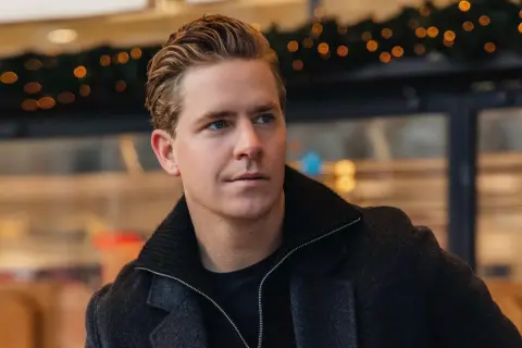 Martin Hansson i stilren höstmode, med en mörk rock och uppsatt hår. Bakgrunden har julbelysning.