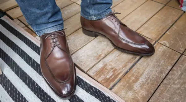 Ett par eleganta bruna läderskor med snörning, som sitter på en person med blå jeans. Skorna har en fin glans och ser ut att vara av hög kvalitet, trots att de har lite glappande skor-känsla.