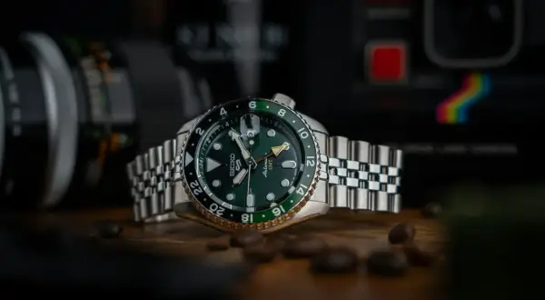 En grön Seiko GMT-klocka med en tvåfärgad bezel, omgiven av kaffebönor. Bakgrunden visar en kamera.