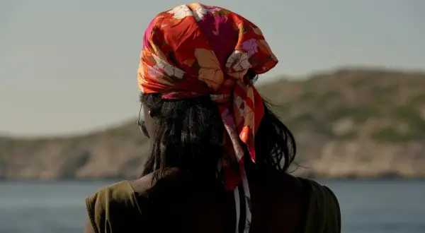 En kvinna med färgglad bandana i håret, en stil som visar hur knyter man en bandana. Bakgrunden är en lugn sjö och berg.