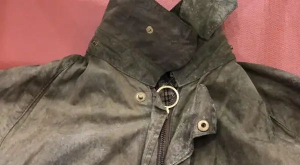 Detaljerad bild av en sliten Barbour-jacka, redo att laga. Kragen är uppfälld och visar dragkedjan med en gyllene ring.