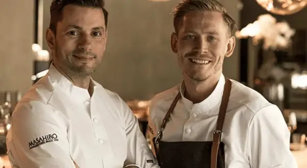 Två kockar ler mot kameran. Deras vita kockrockar och förkläden antyder en professionell miljö, kanske en restaurang som får bra allegrine recensioner.