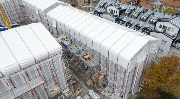 Byggarbetsplats i Sverige med vita presenningar över hus, ställningar och en gul kran. En pampuscher herr skulle uppskatta den ordning och reda som råder.