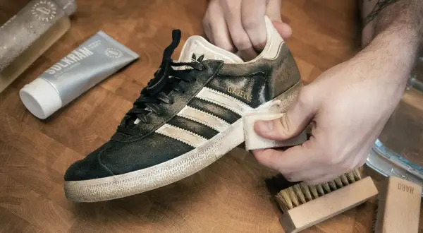 Händer rengör en smutsig Adidas Gazelle-sneaker med en trasa. Bredvid ligger rengöringspasta och borstar, redo att impregnera sneakers.