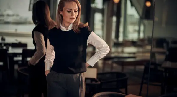 En kvinna med blont hår och fräknar bär en vit skjorta och en svart väst. Hon visar hur en frack ska sitta, med en avslappnad men elegant hållning.