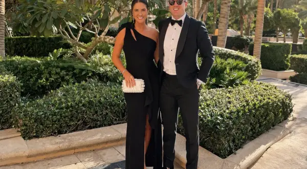Ett par klädda för black tie-tema, hon i en elegant svart klänning, han i smoking. De poserar glatt utomhus bland grönska.