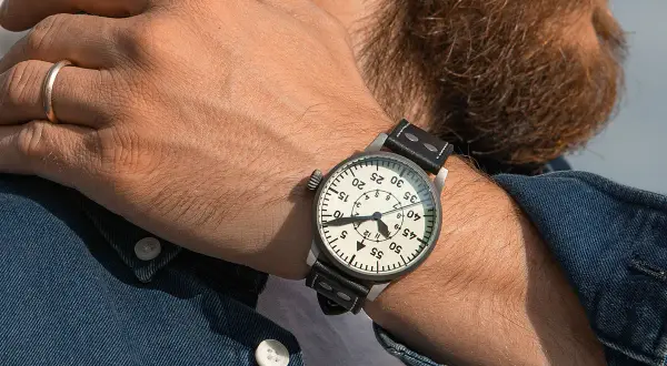En man med skägg bär en klassisk fliegerklocka med tydlig urtavla och läderarmband.