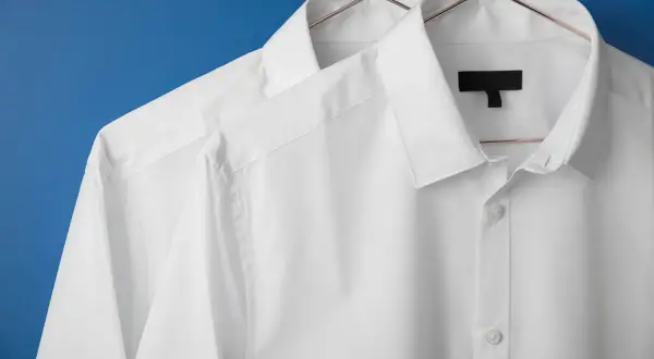 Två vita skjortor hänger på galgar mot en blå bakgrund. De ser ut som om de precis kommit från en **dry cleaner**, perfekt strukna och redo att bäras.