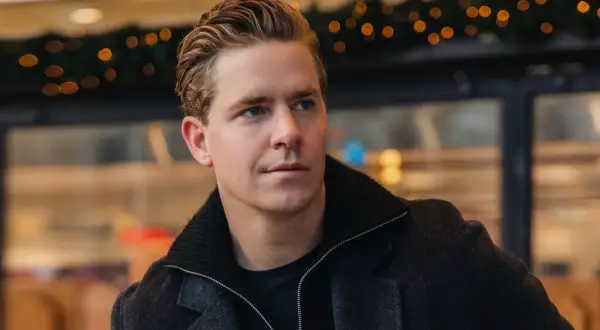 Martin Hansson i stilren höstmode, med en mörk rock och uppsatt hår. Bakgrunden har julbelysning.