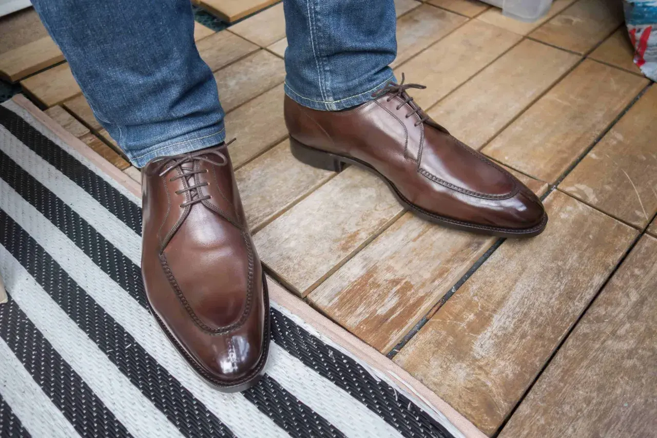 Ett par eleganta bruna läderskor med snörning, som sitter på en person med blå jeans. Skorna har en fin glans och ser ut att vara av hög kvalitet, trots att de har lite glappande skor-känsla.