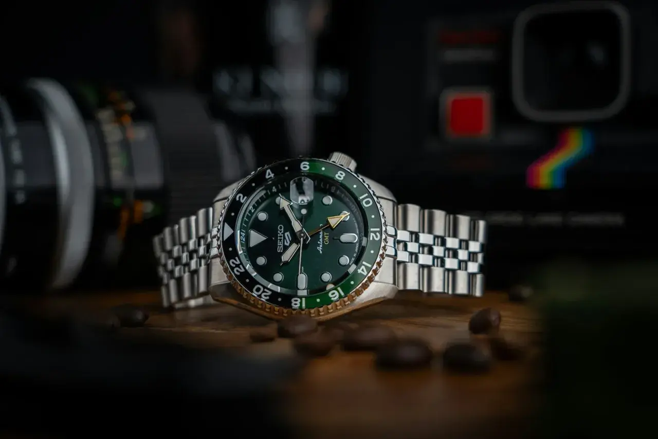 En grön Seiko GMT-klocka med en tvåfärgad bezel, omgiven av kaffebönor. Bakgrunden visar en kamera.