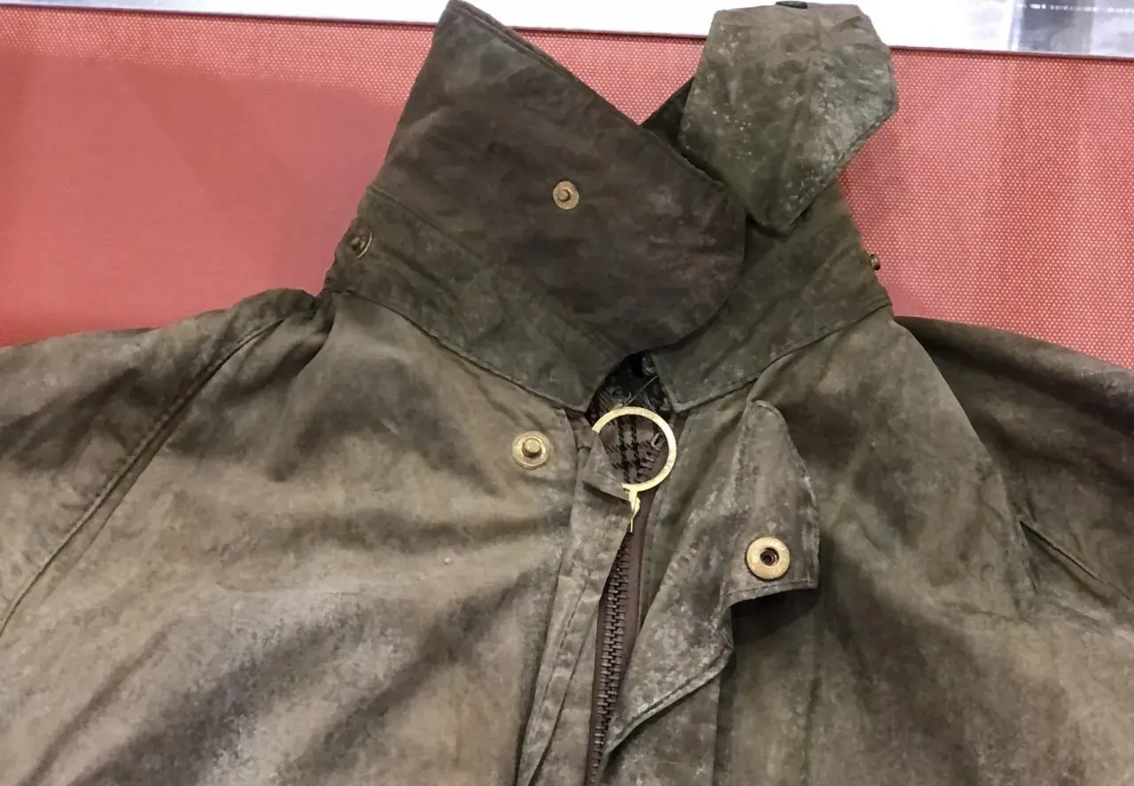 Detaljerad bild av en sliten Barbour-jacka, redo att laga. Kragen är uppfälld och visar dragkedjan med en gyllene ring.