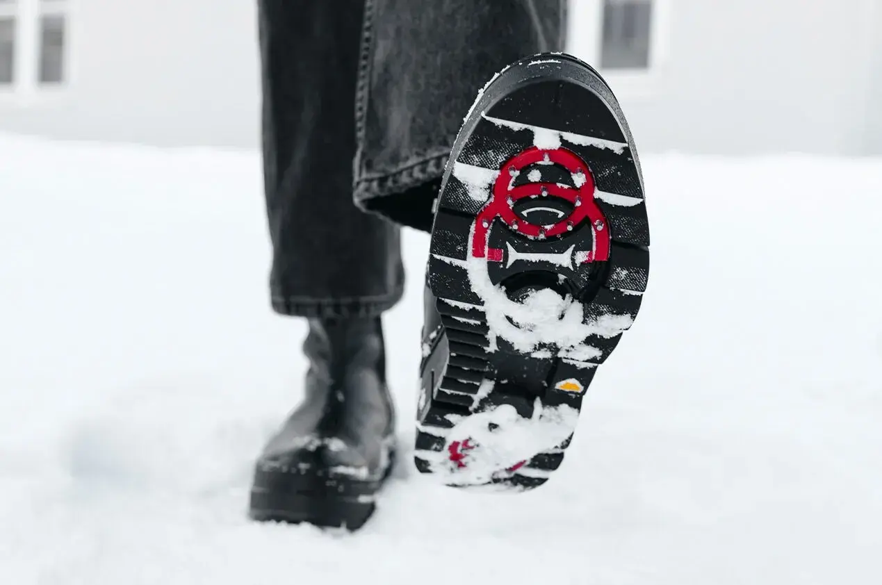 En person går i snön med svarta stövlar. Sulorna har ett mönster och röda detaljer för att undvika hala skor.