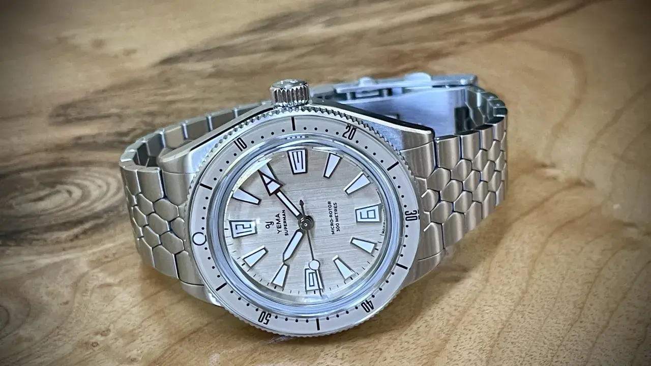 En Yema Superman dykarklocka med silverfärgad urtavla och ett unikt, bikakeformat metallarmband ligger på ett träbord.
