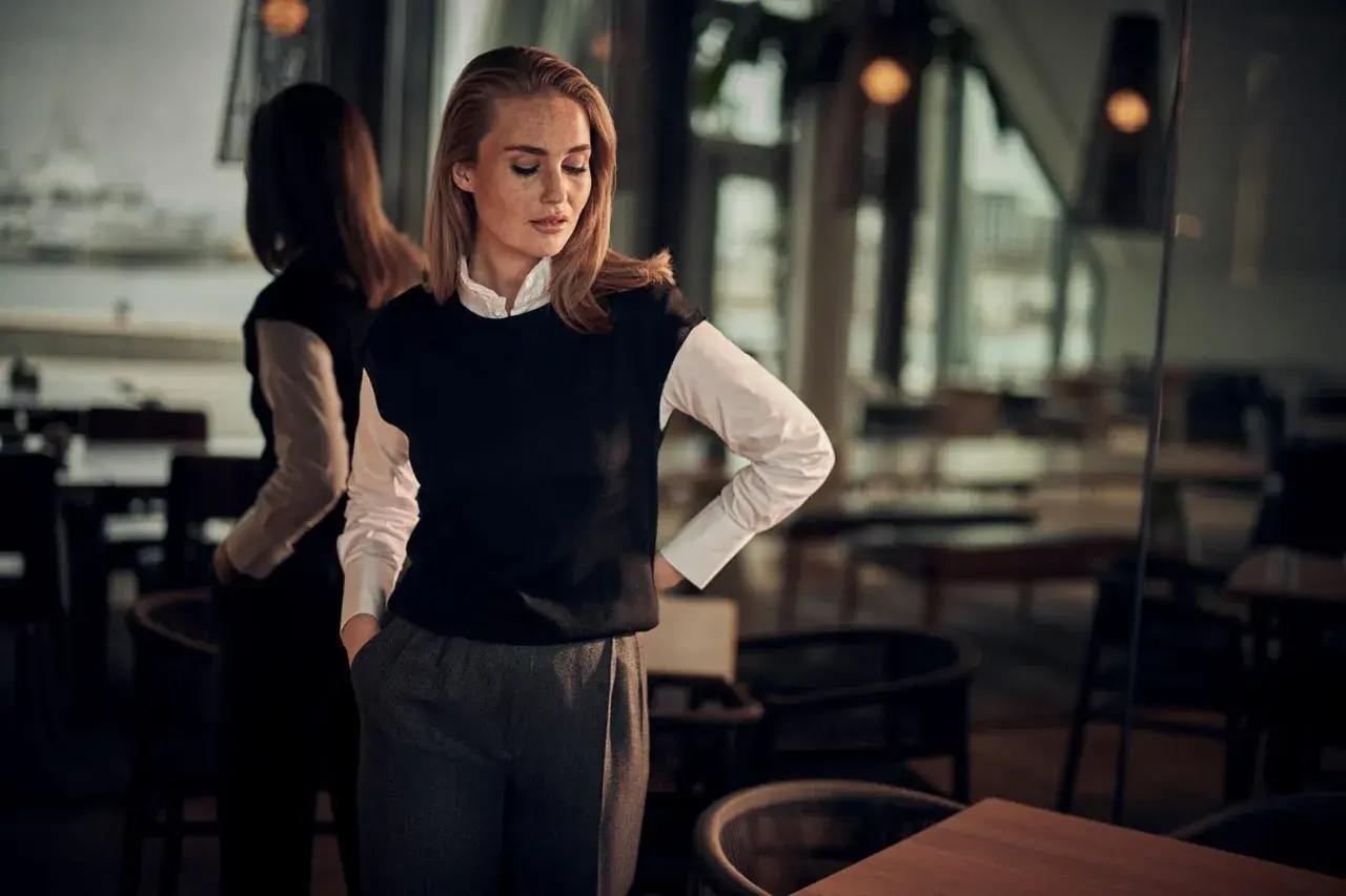 En kvinna med blont hår och fräknar bär en vit skjorta och en svart väst. Hon visar hur en frack ska sitta, med en avslappnad men elegant hållning.