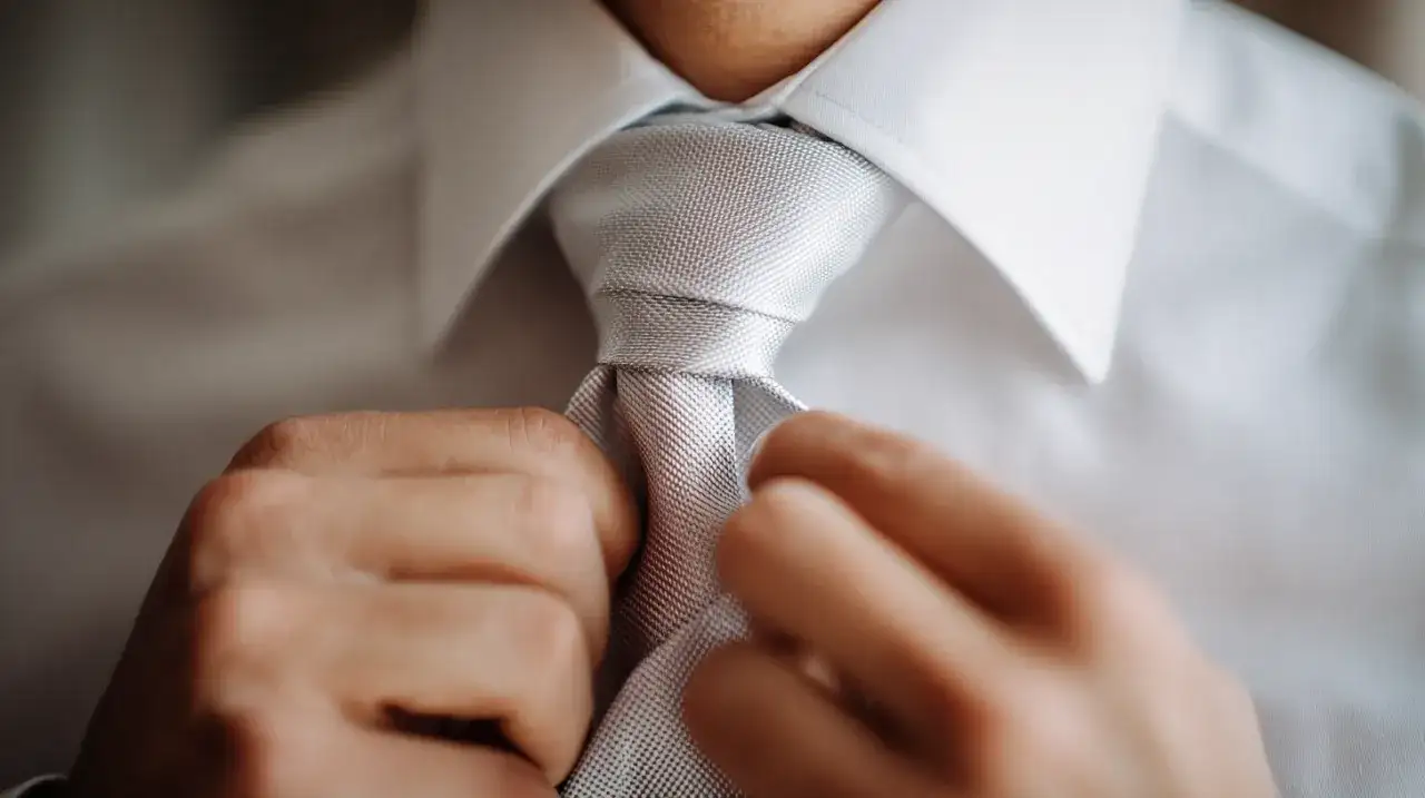 Händer som knyter en ljusgrå slips till en vit skjorta. En viktig detalj för att knyta slipsen perfekt.
