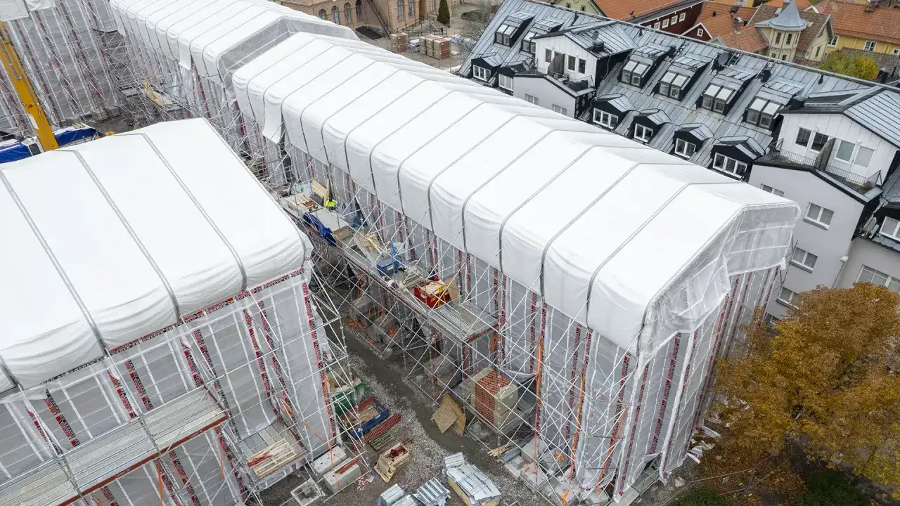 Byggarbetsplats i Sverige med vita presenningar över hus, ställningar och en gul kran. En pampuscher herr skulle uppskatta den ordning och reda som råder.