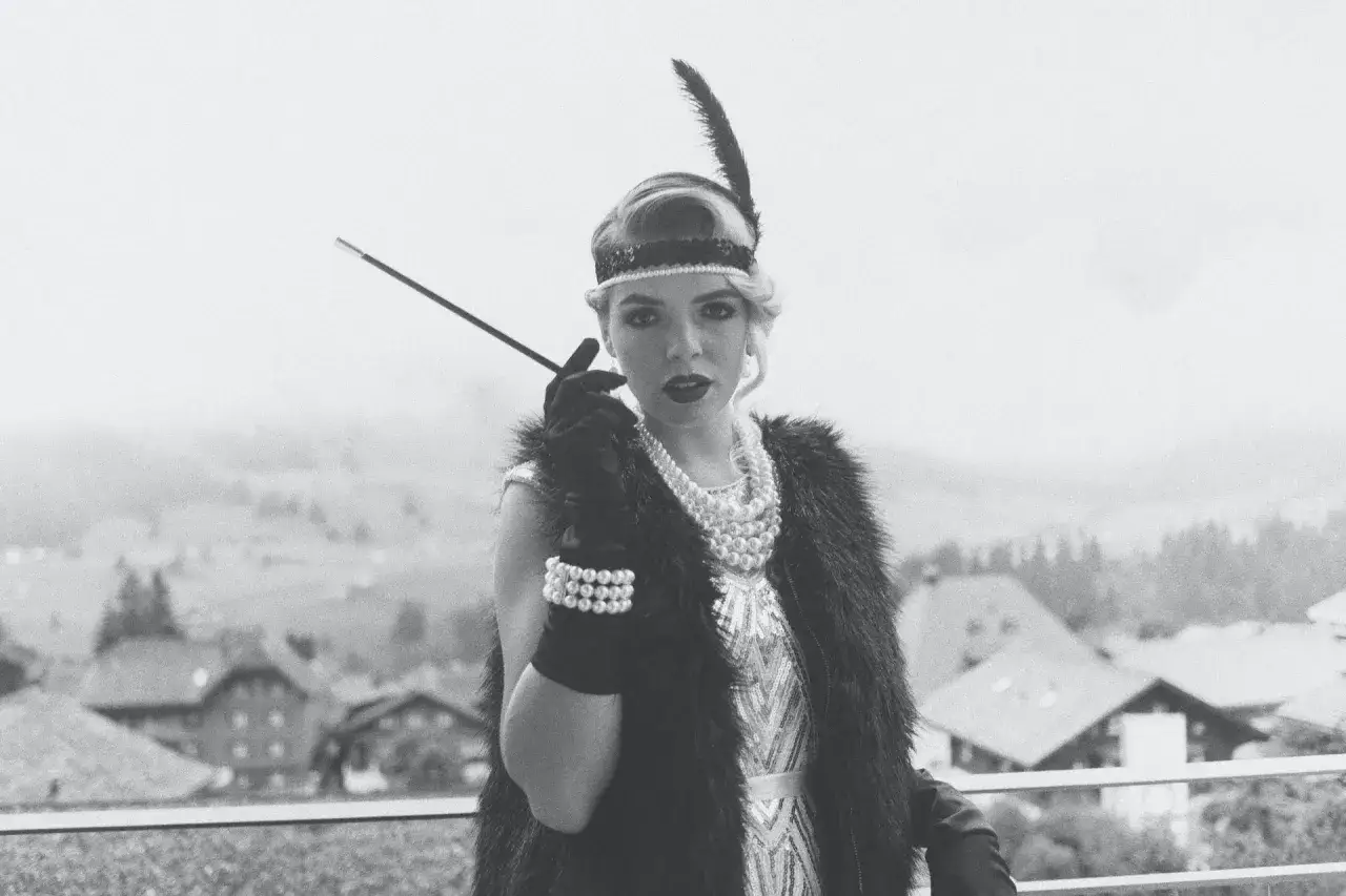 En kvinna i mode 1920-tal med fj&auml;derpannband och p&auml;rlhalsband h&aring;ller en cigarettmunstycke.
