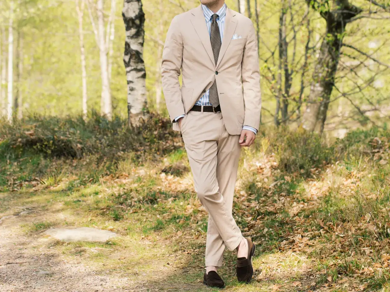 Man i ljusbeige kostym med slips och mockasiner, stående i en skogsmiljö.