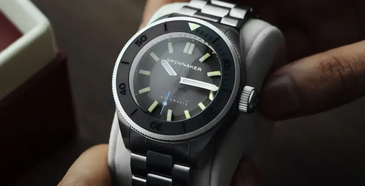 En nära bild av en Spinnaker klocka med mörk urtavla och lysande index. Klockan har en dykarring och ett metallarmband.