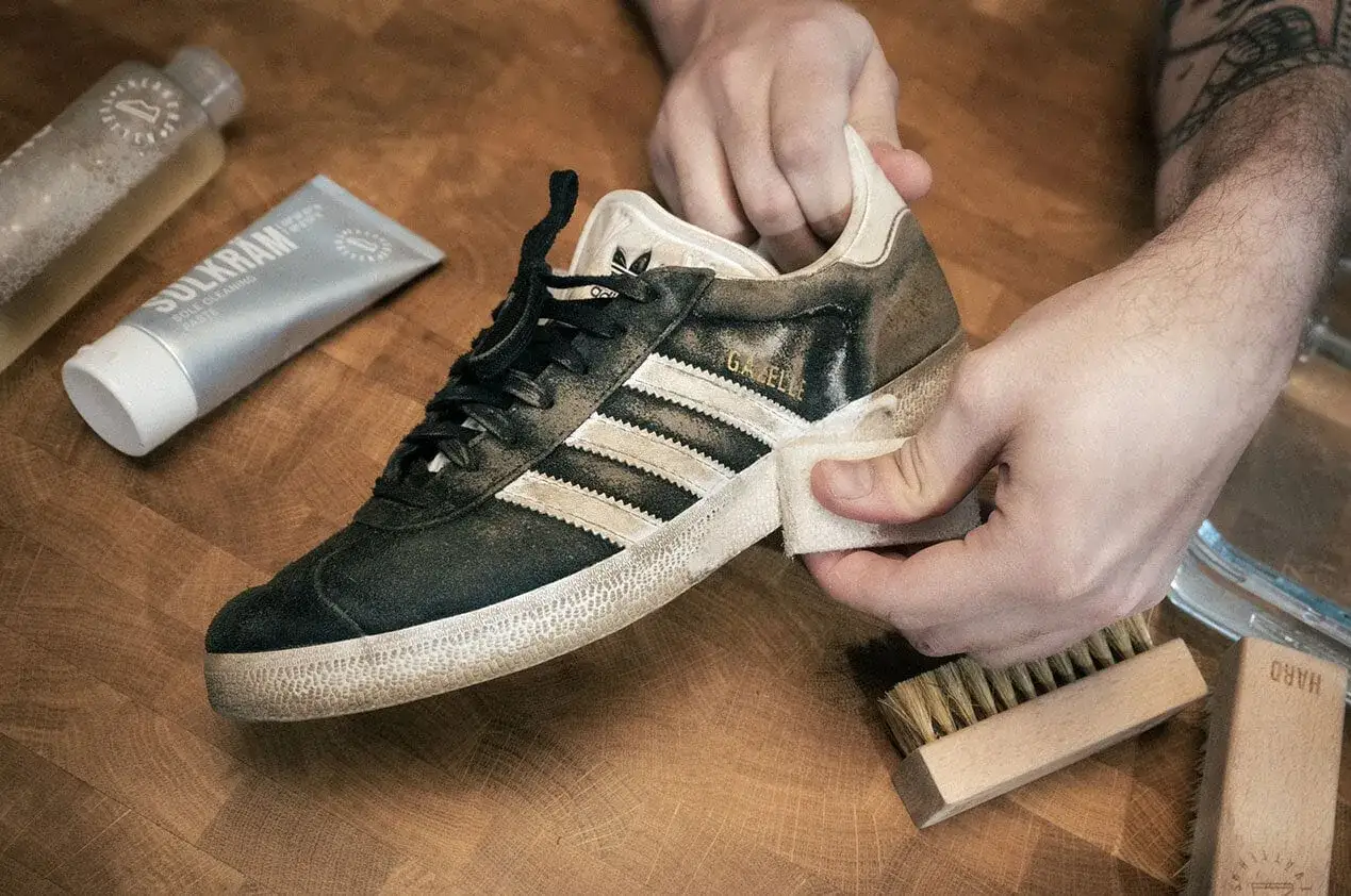 Händer rengör en smutsig Adidas Gazelle-sneaker med en trasa. Bredvid ligger rengöringspasta och borstar, redo att impregnera sneakers.
