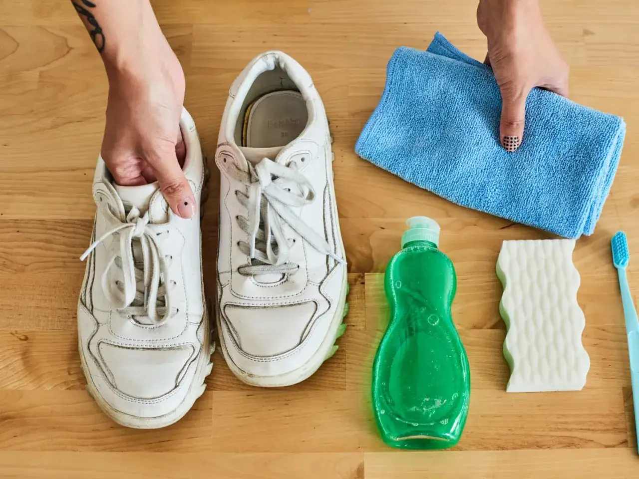 Händer som håller ett par smutsiga vita sneakers bredvid rengöringstillbehör. Målet är att få vita skor vita igen.
