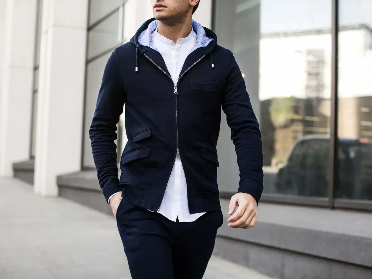 Sebastian Dollinger i mörkblå hoodie och vit skjorta, med handen i fickan, går längs en byggnad.