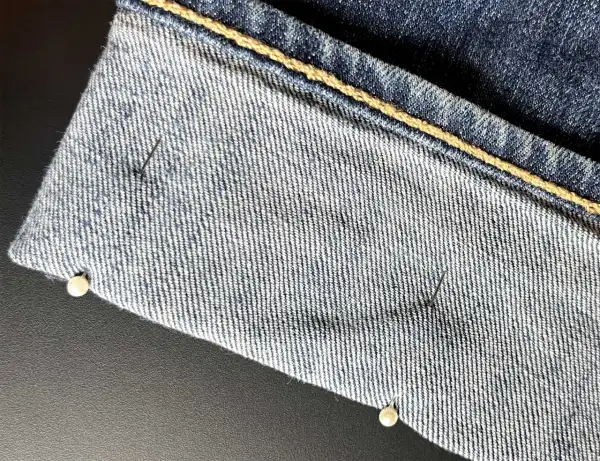 Närbild på jeans som ska vikas upp. Nålar håller tyget på plats.