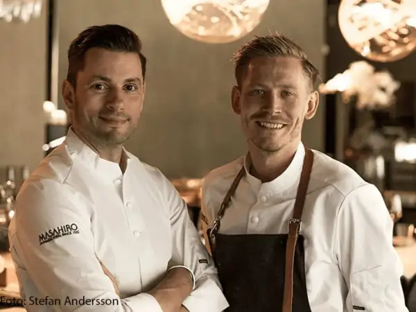 Två kockar ler mot kameran. Deras vita kockrockar och förkläden antyder en professionell miljö, kanske en restaurang som får bra allegrine recensioner.