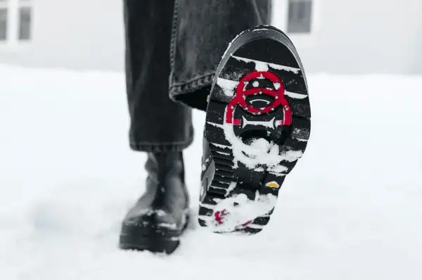 En person går i snön med svarta stövlar. Sulorna har ett mönster och röda detaljer för att undvika hala skor.